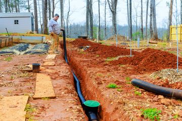 Trenched red clay yard with black pipes and a worker near a house under construction
