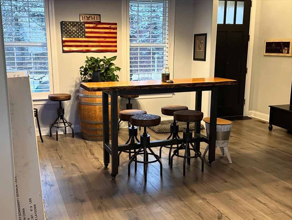 Room with bar table, stools, wooden flooring, American flag art, and a barrel.