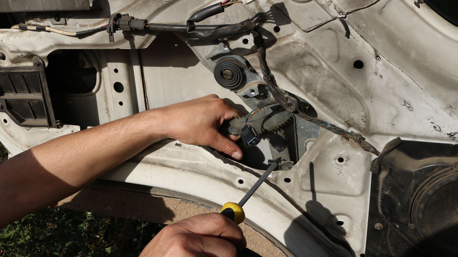 Hands using a screwdriver to adjust a car door window mechanism inside the exposed door frame.