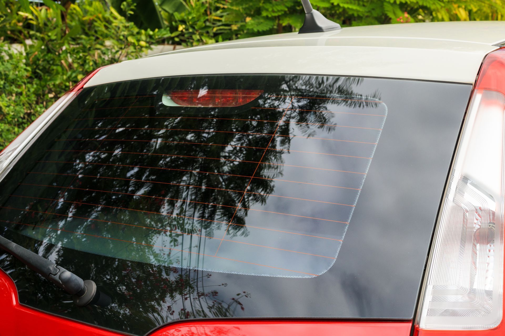Rear window of a red car with a dark tinted hatch glass reflecting trees and sky