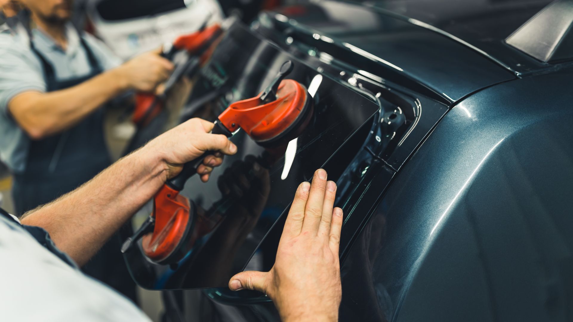 Two hands use red suction cups to remove a glossy dark car panel in a workshop.