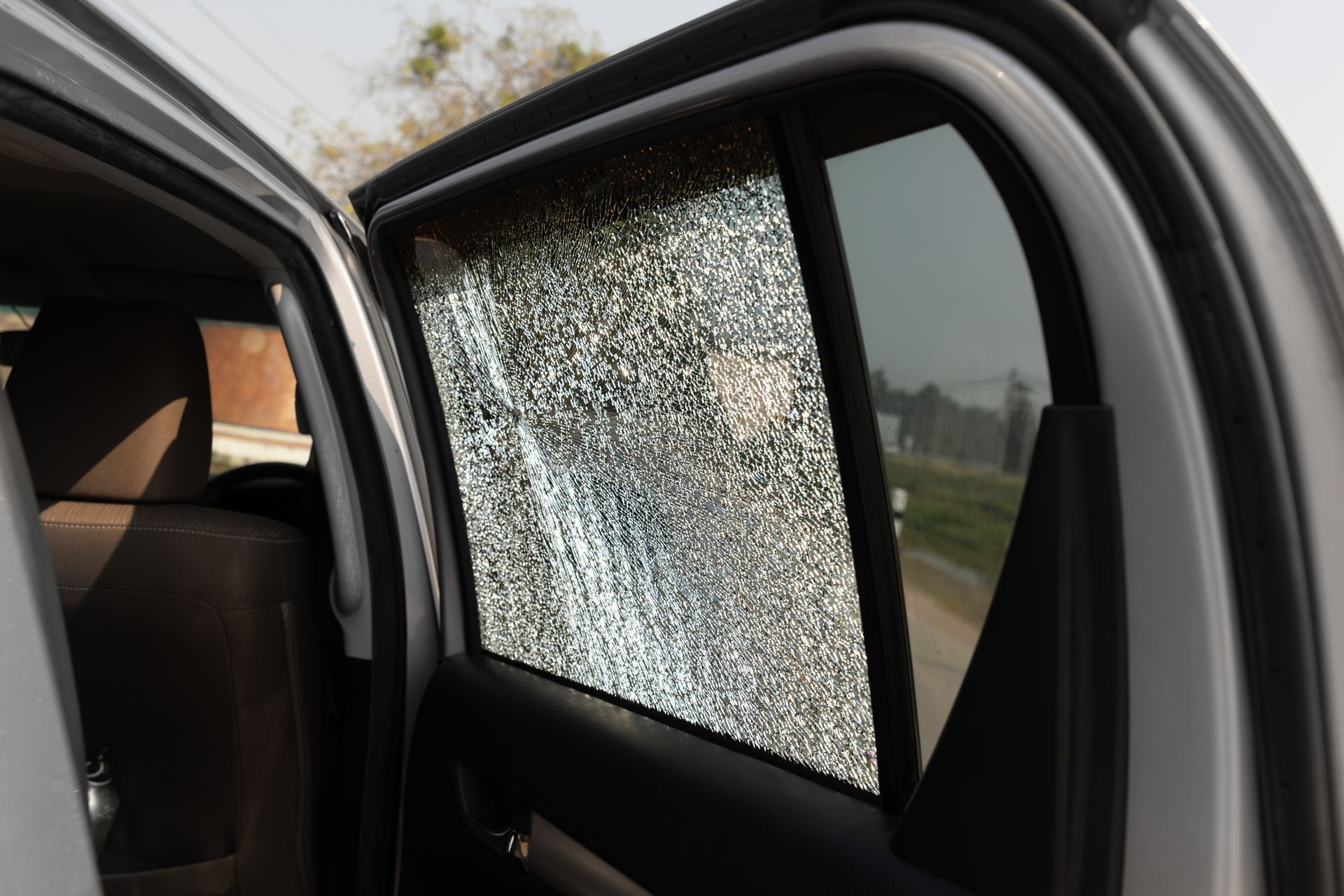 Car side window shattered with safety glass cracked, viewed from outside on a roadside.