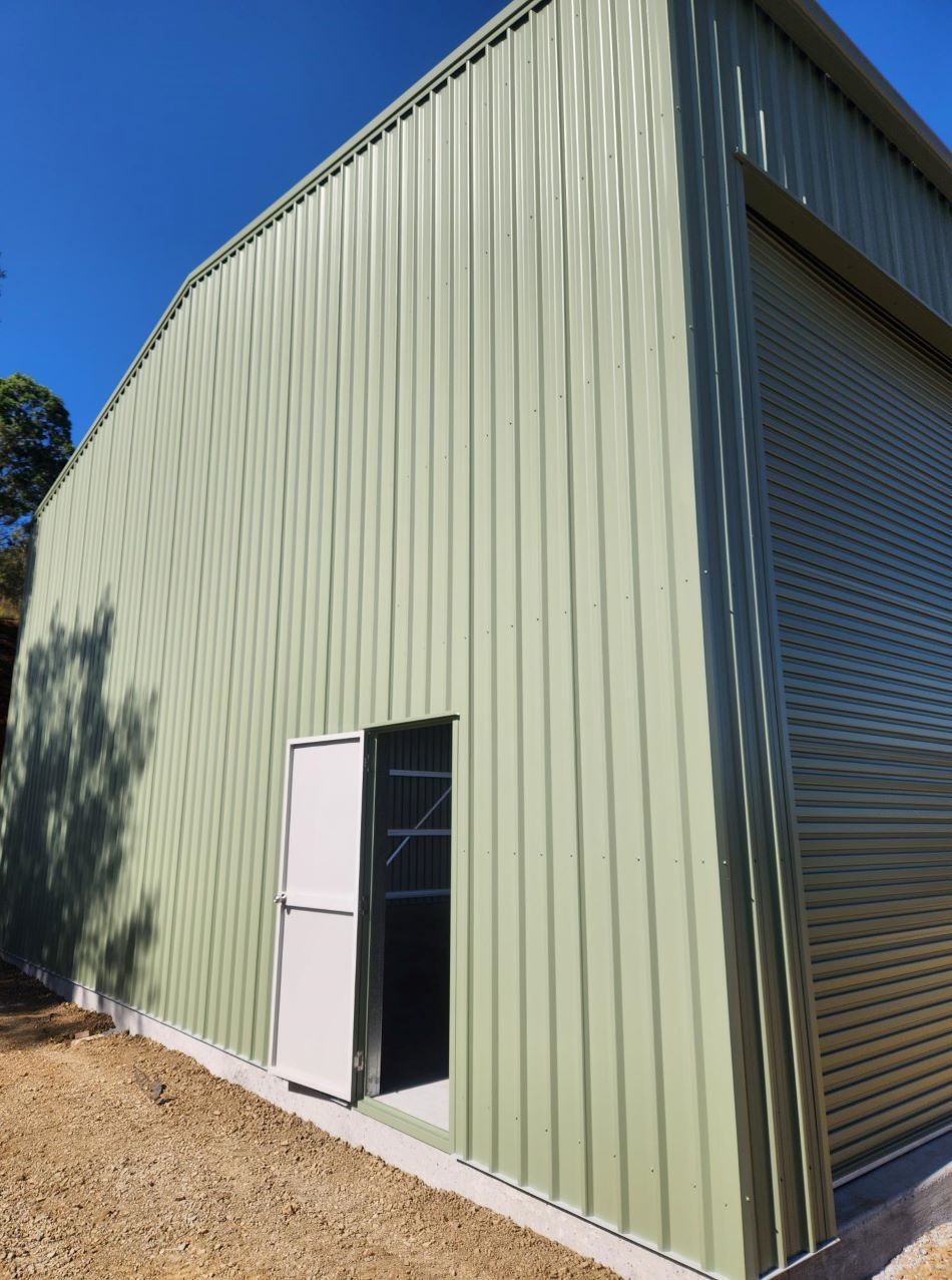 A Green Metal Building With a White Door and Roller Shutters — Whit Coast Sheds in  Gregory River, QLD