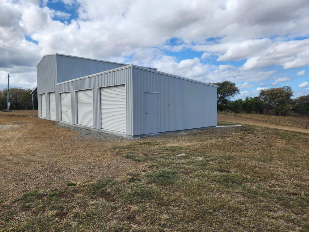Gray metal storage building with four garage doors on a grassy lot under cloudy skies— Whit Coast Sheds in Gregory River, QLD