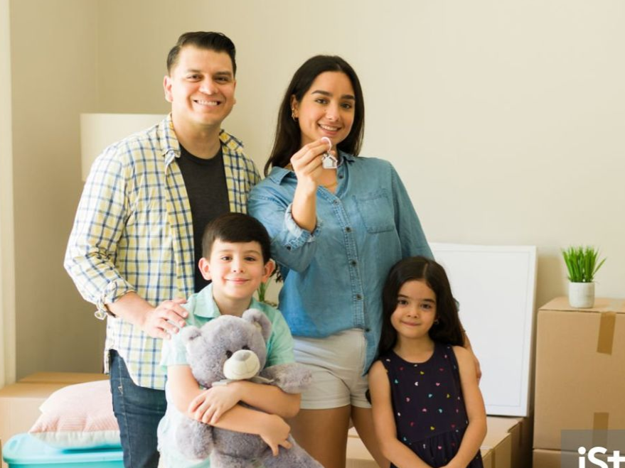 Family of four smiles, woman holds house keys; in new home with moving boxes.