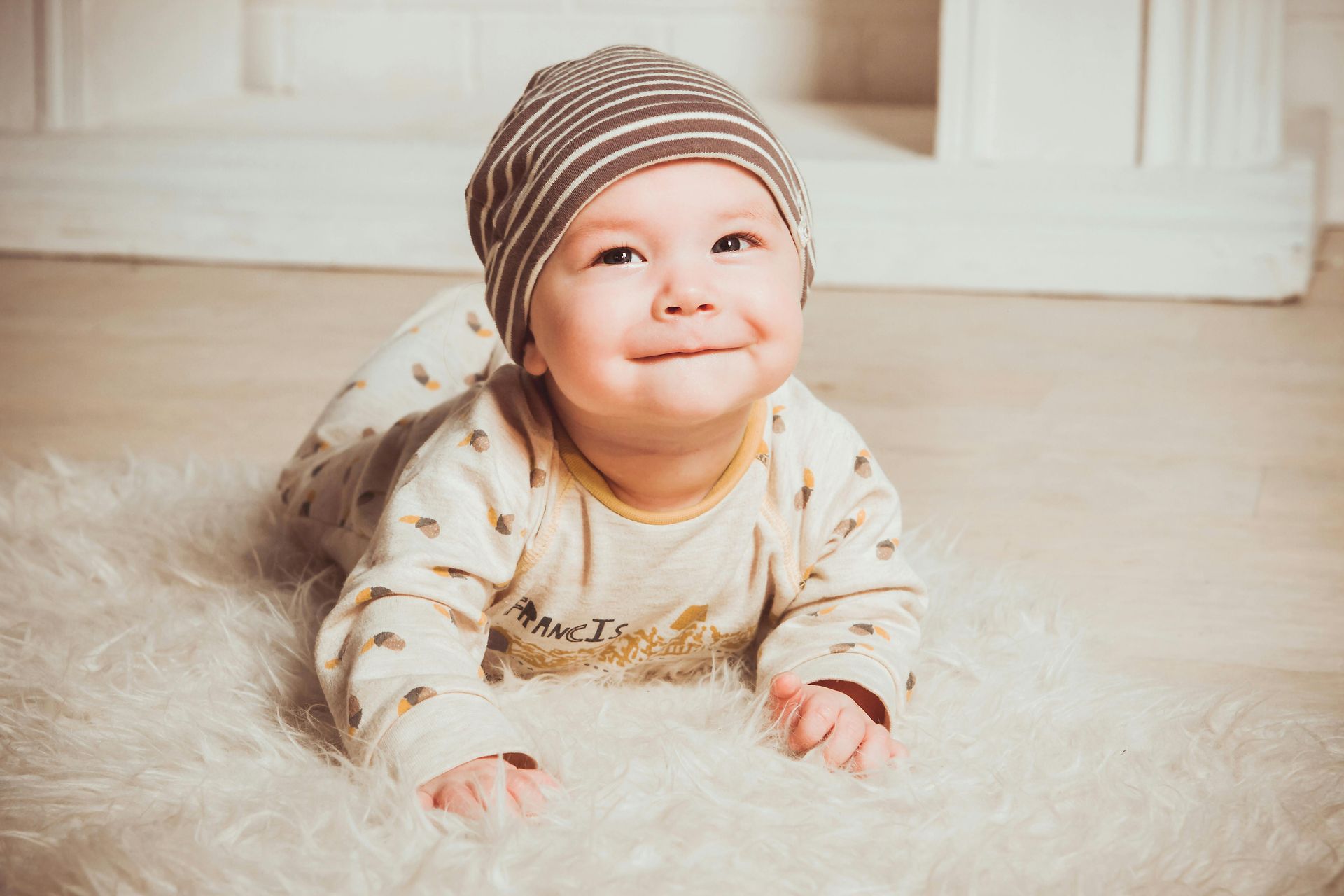 Baby wearing a knit hat, smiling, on a white fluffy rug.
