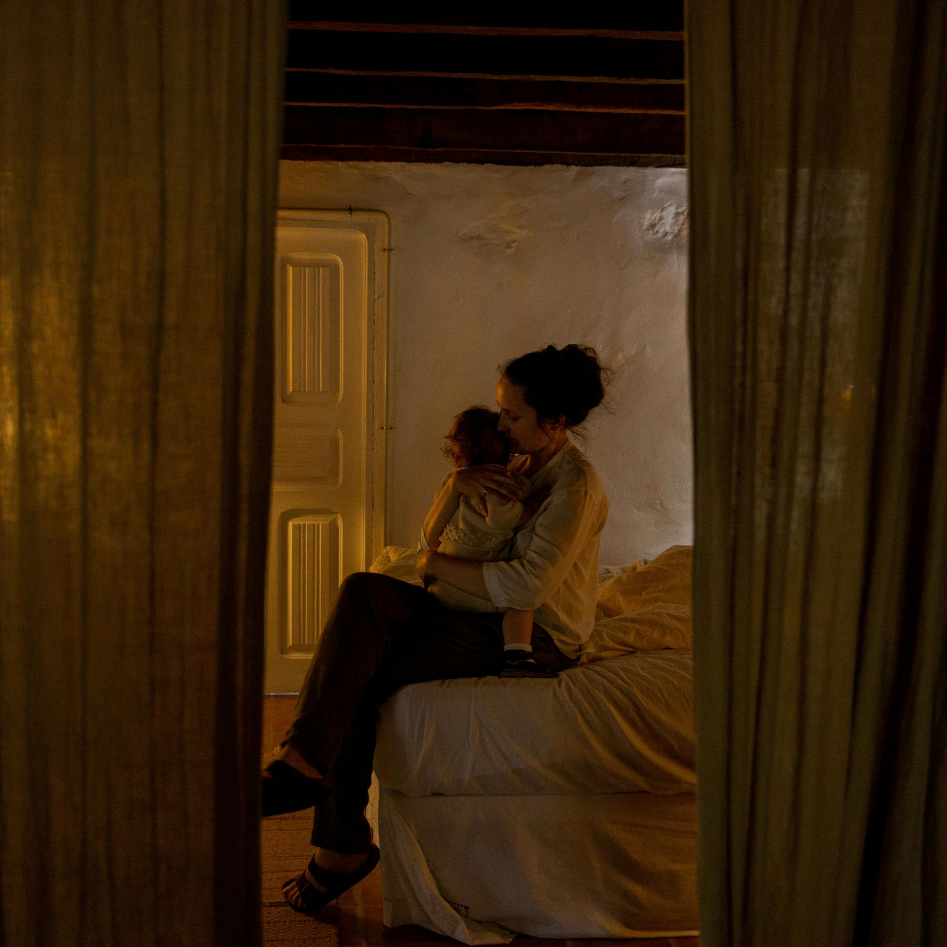 Woman holding a baby sits on a bed, viewed through curtains, in a dimly lit room.