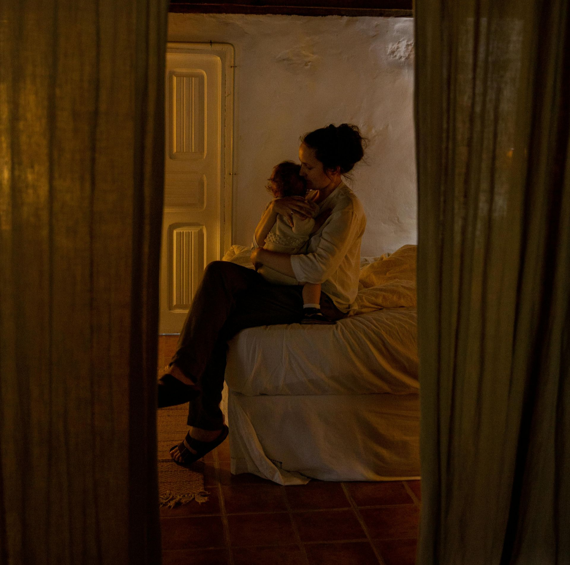 Woman seated on bed, holding a baby. Warm lighting, partially visible room.