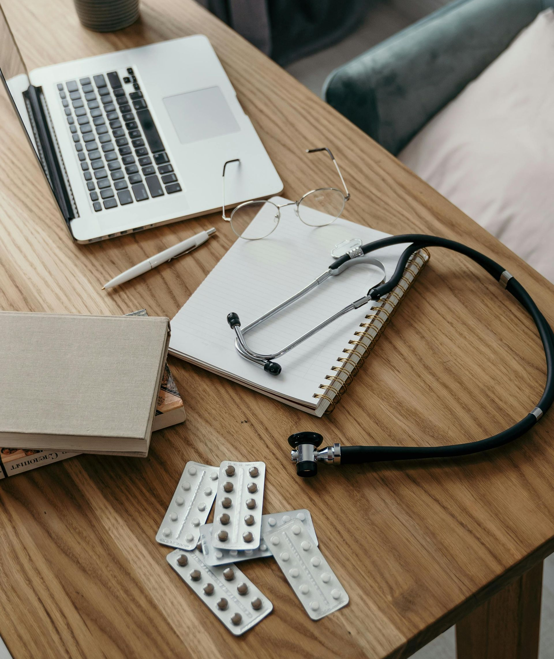 Laptop, stethoscope, notebook, pen, and pills on a wooden table.