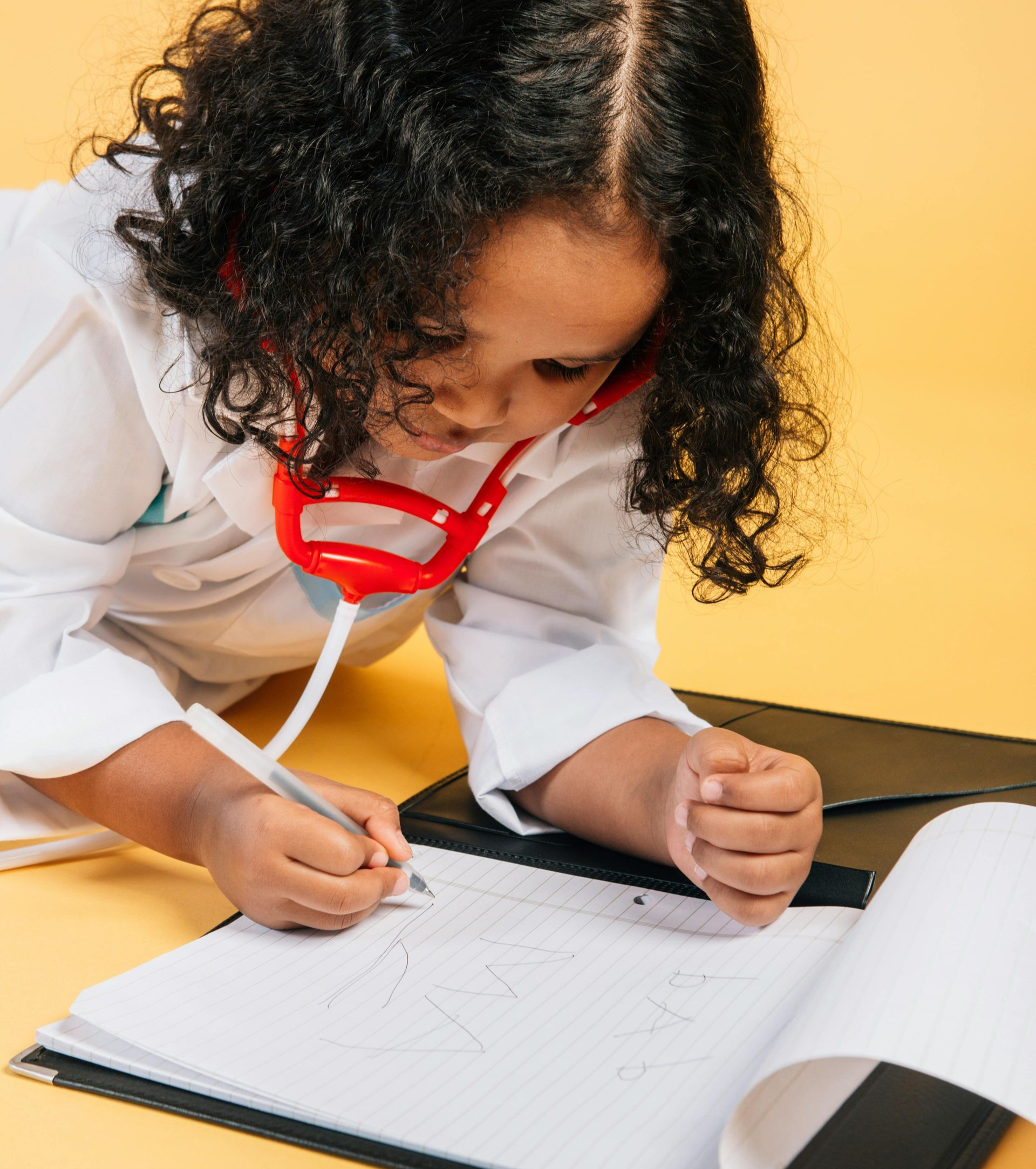 Child wearing a lab coat and stethoscope, writing in a notebook on a yellow surface.