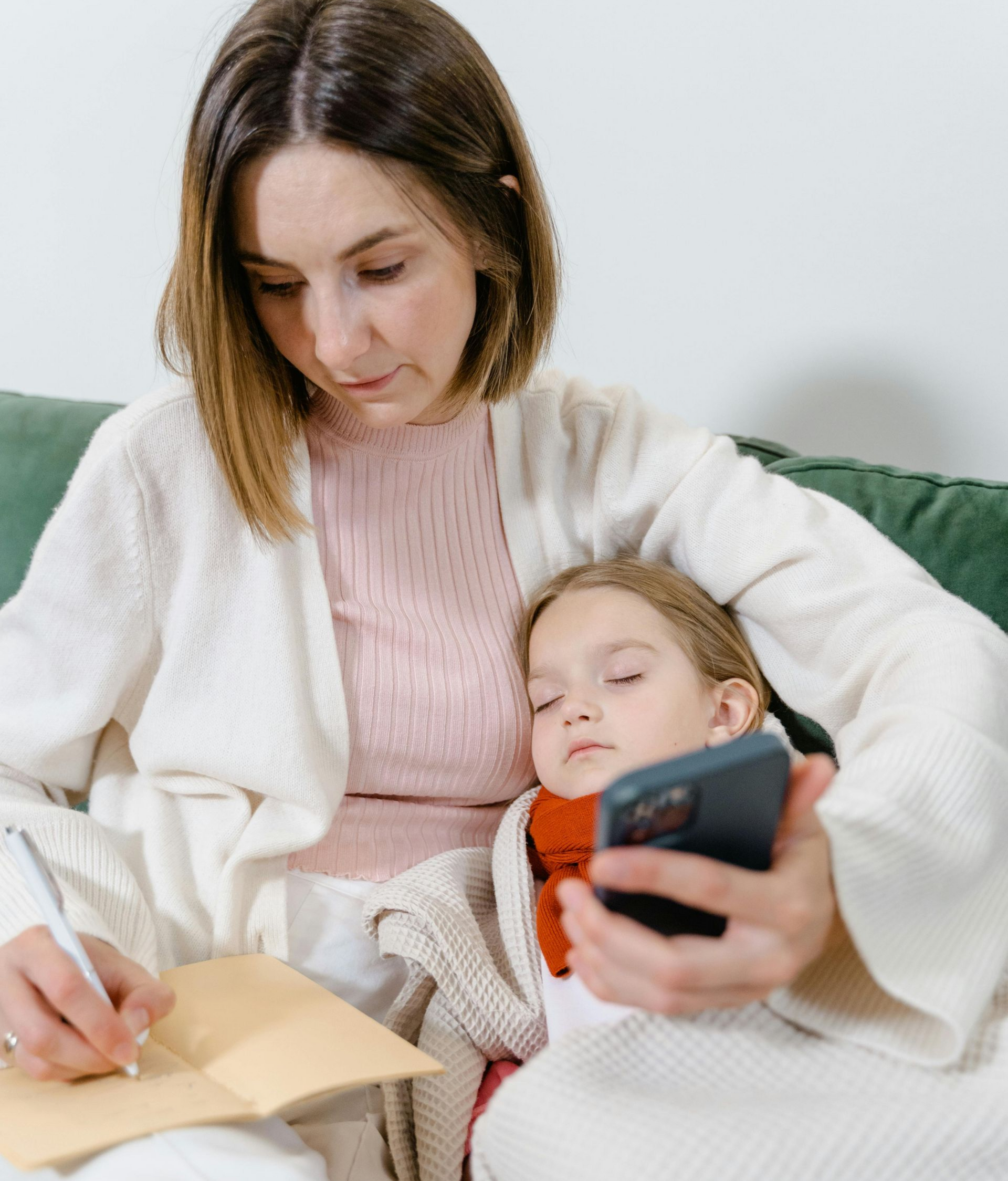 Woman writing in notebook while holding phone and child resting on her lap.