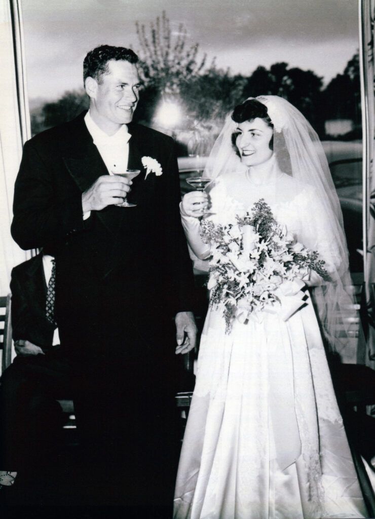 Wedding couple toasting. Man in tuxedo, woman in white gown holding bouquet, both smiling. Restaurant setting.