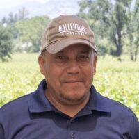 Man in a cap and blue shirt smiling outdoors in front of a vineyard.