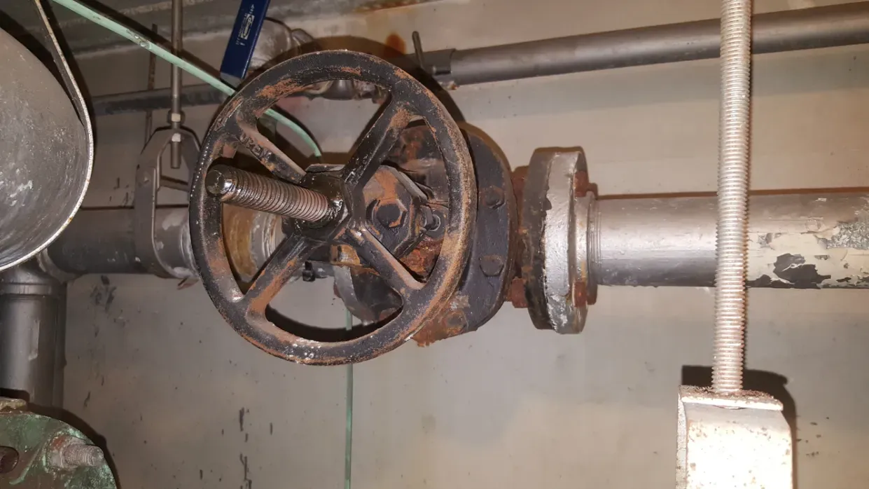 Industrial pipe with a large rusted valve wheel mounted on a metal pipeline in a utility room