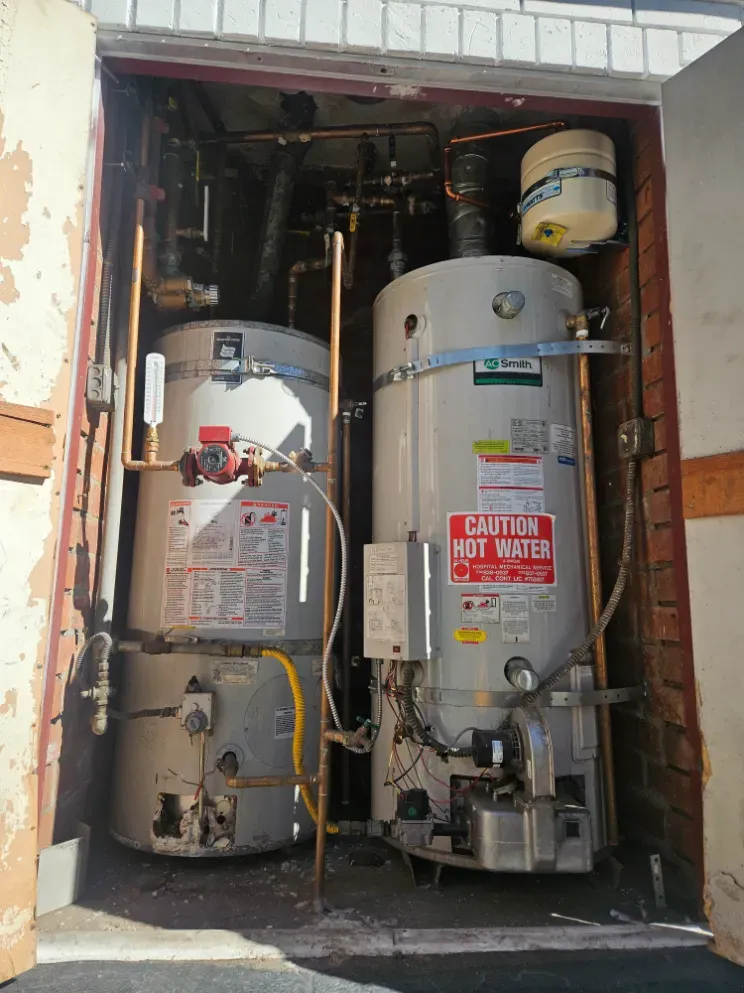 Two water heaters in a utility closet, with pipes, valves, and a warning sign.