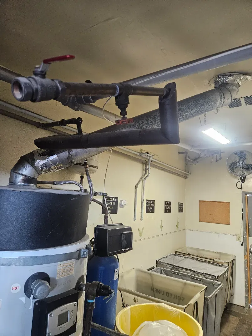 Boiler room with large pipes, tanks, and utility equipment, plus a yellow bucket on the floor.