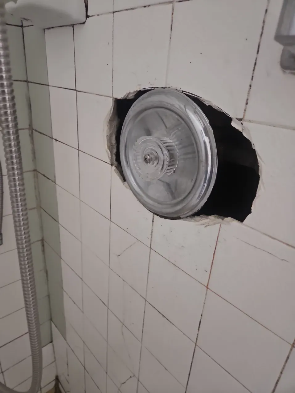 Broken bathroom wall tiles around a round metal vent or fan cover