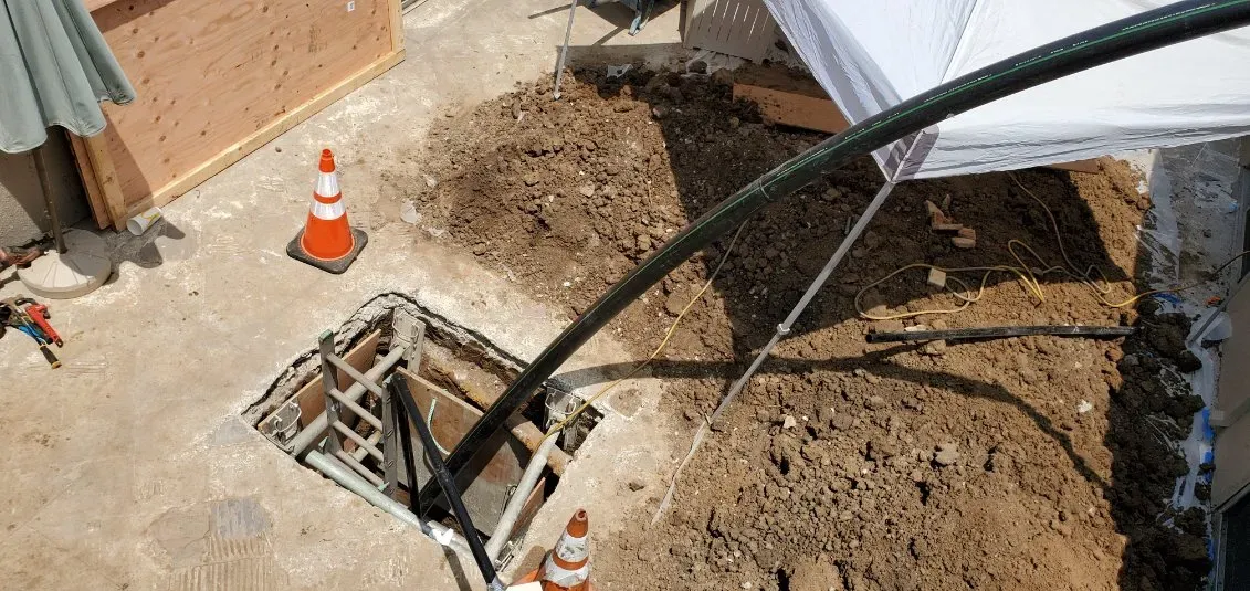 Construction trench beside a building with dirt pile, caution cone, and a metal ladder