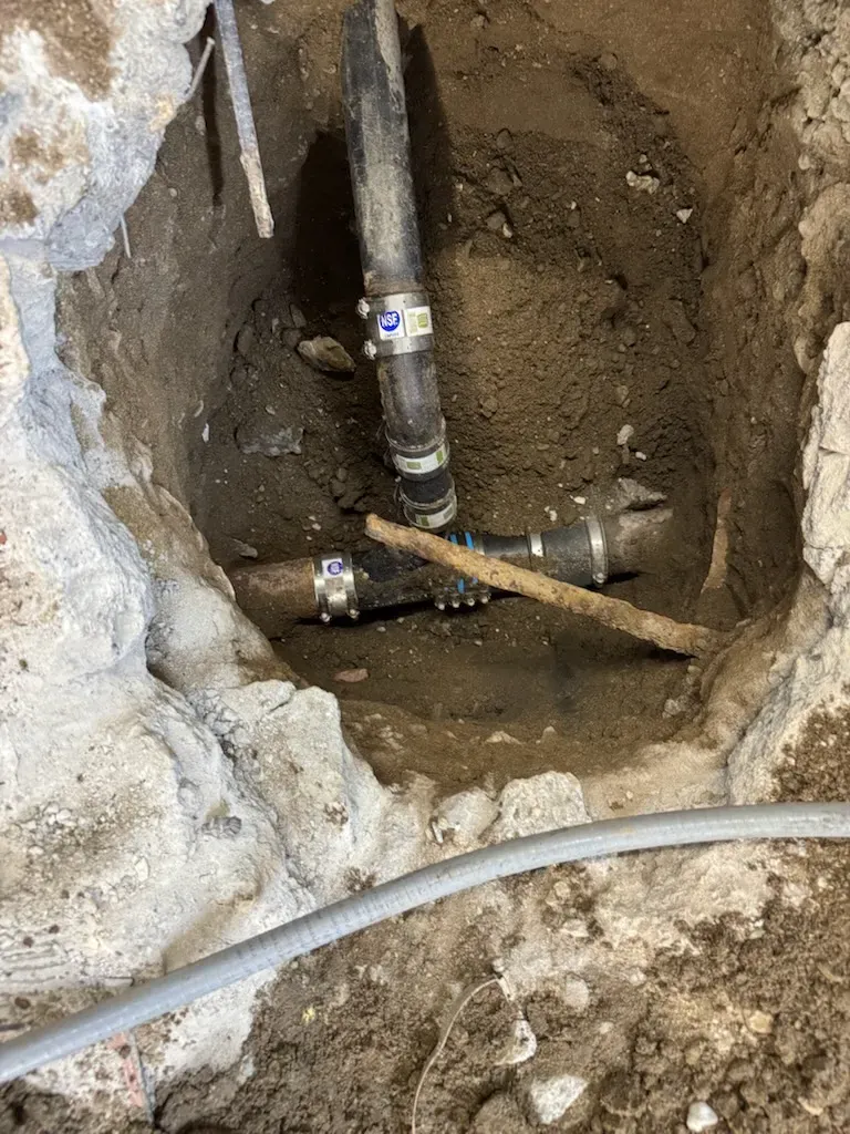 Exposed plumbing pipes in a dirt wall excavation, with a flexible hose across the trench.