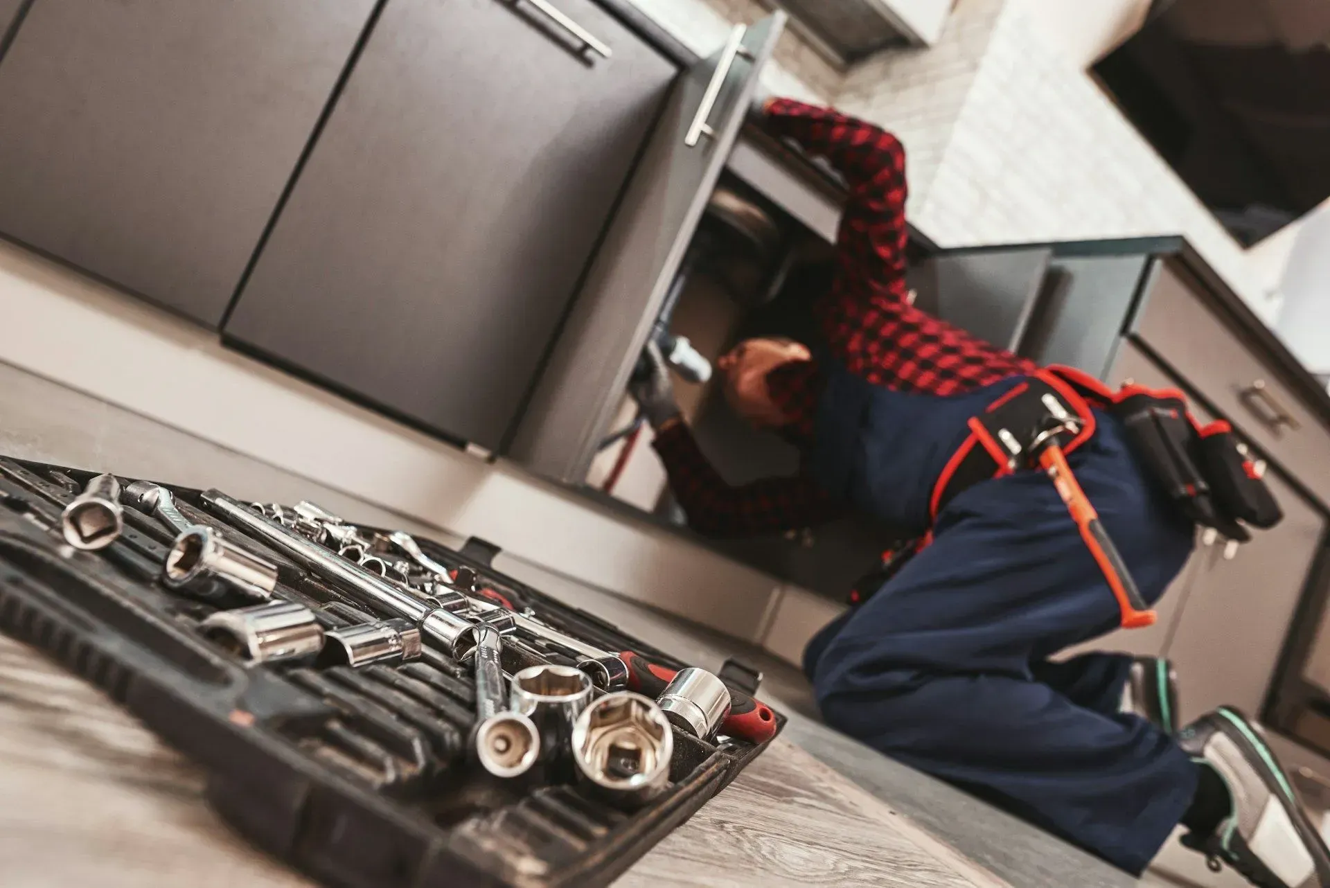 Mechanic kneeling under a car, using tools; toolbox in foreground, workshop setting