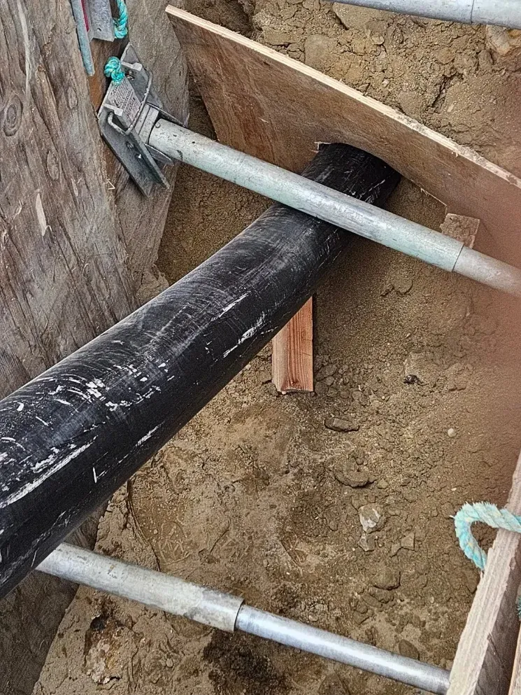 Black pipe through a dirt wall opening, held by metal scaffolding poles in a construction trench