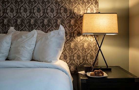 Bed with white pillows beside a lit bedside lamp on a dark nightstand in a patterned bedroom