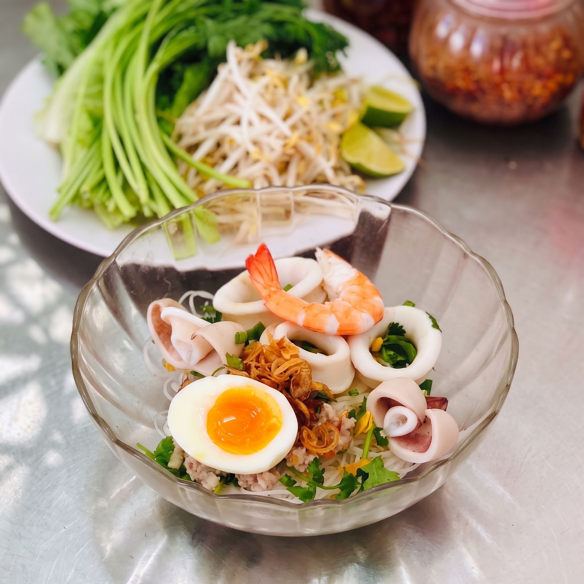 Glass bowl of seafood noodle salad with shrimp, boiled egg, and herbs on a table, with fresh greens behind