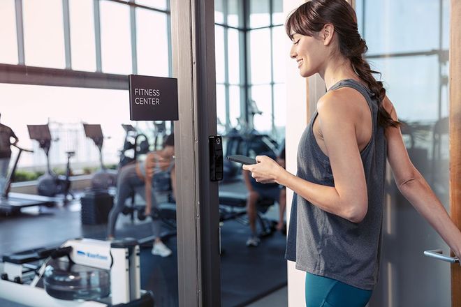 Woman opening the door to a fitness center, holding a phone. People are working out inside.