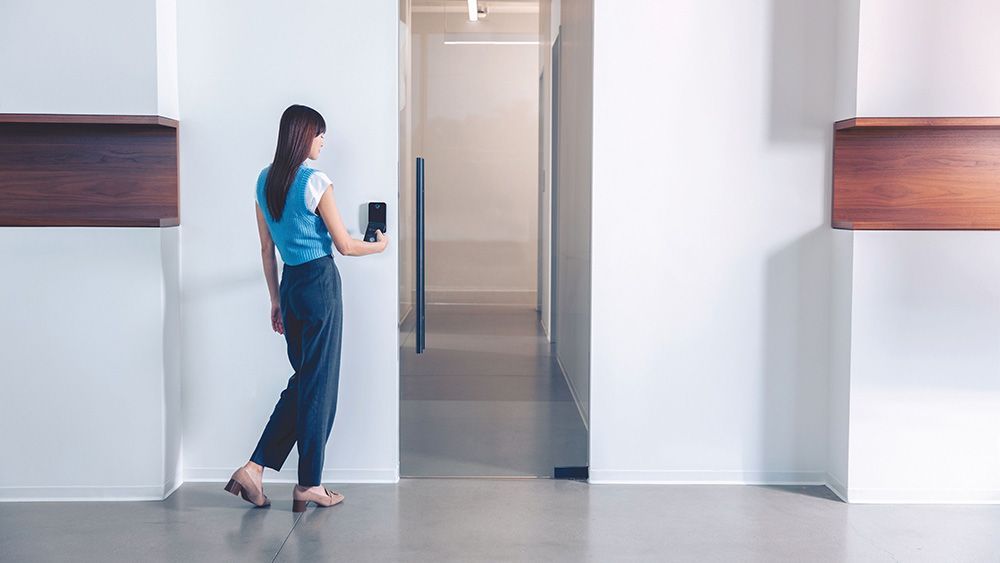 Woman entering a doorway, using a key card reader. The setting is a minimalist white hallway with wooden accents.