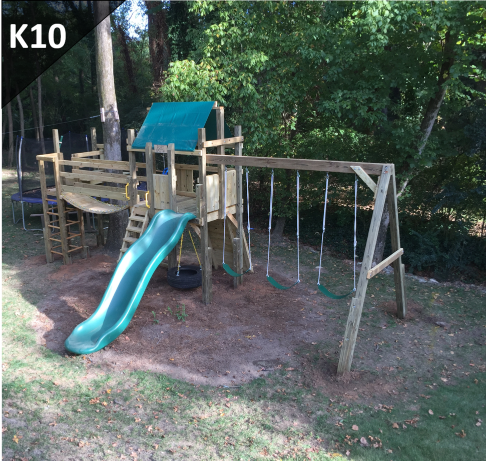 Wooden backyard playset with green slide, swings, and a small tower under trees