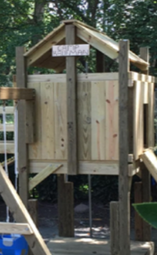 Wooden playhouse under construction with a peaked roof in a wooded outdoor area