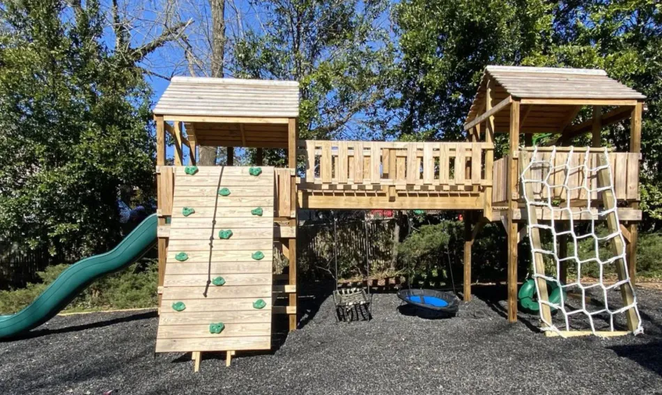 Children’s playground with wooden climbing structures, slide, rope net, and tire swing on gravel.