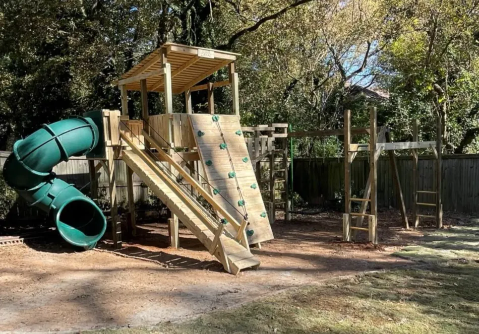 Wooden playground tower with green tube slides and climbing ramp in a shaded park