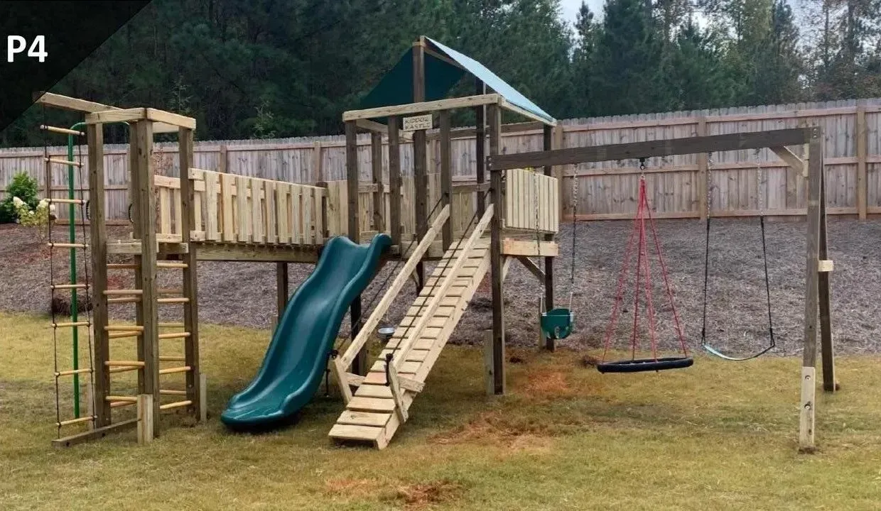 Wooden playground with green slides, ladder, rope swing, and climbing wall in a grassy yard.