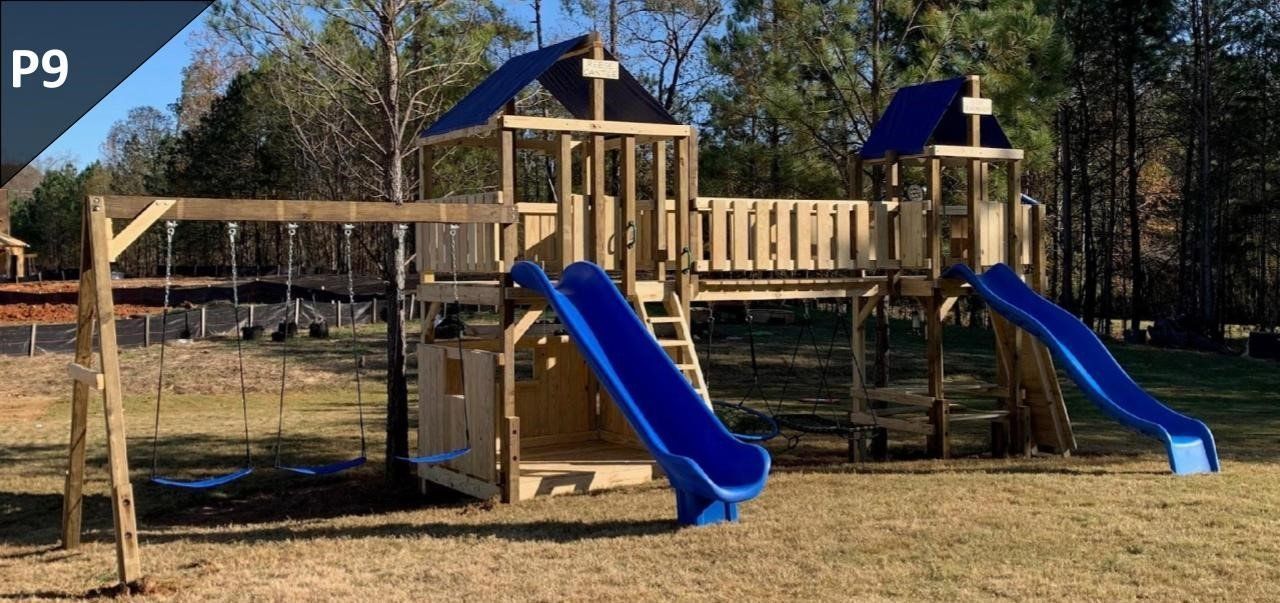 Playground with two blue slides, wooden climbing structure, and swing set in a grassy park area.