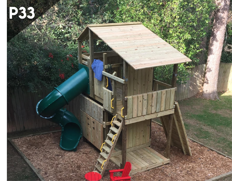 Wooden backyard playhouse with green slide, ladder, and small chairs under trees.