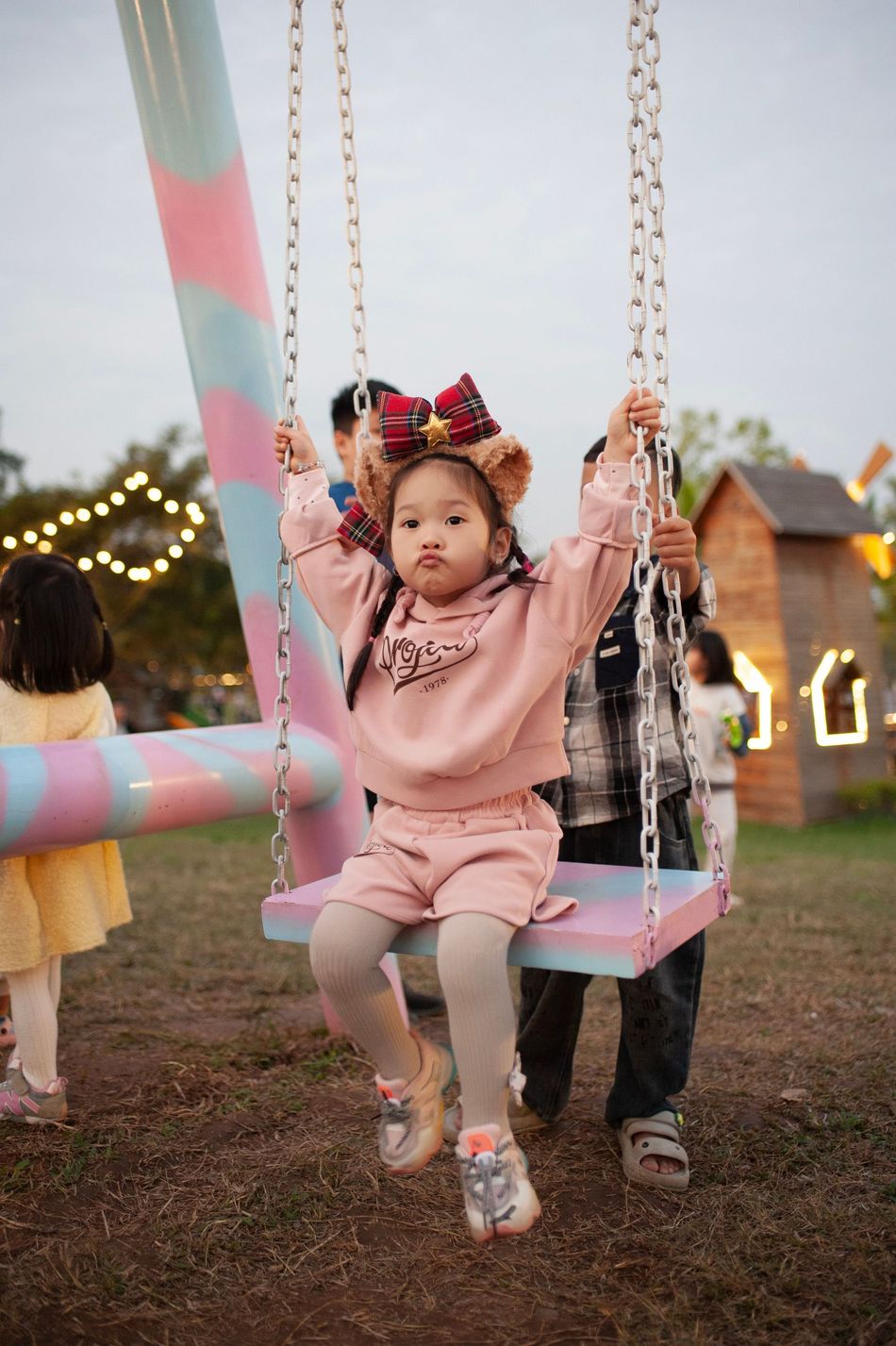 Child on a swing at an outdoor fair, wearing a pink outfit and floral headband, with lights in the background.