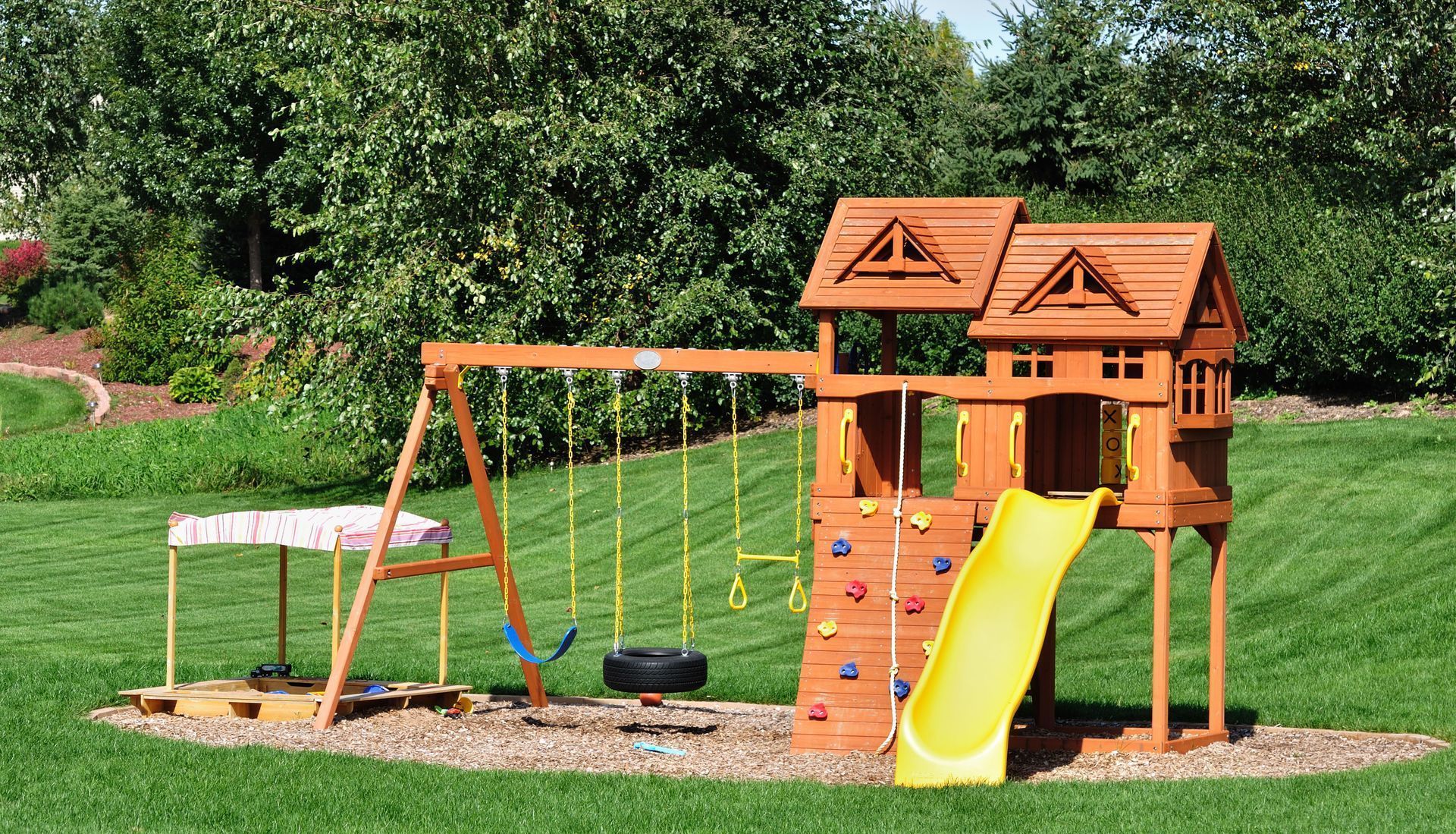 Children's playground with wooden playset, yellow slide, swings, and a small climber in a grassy park