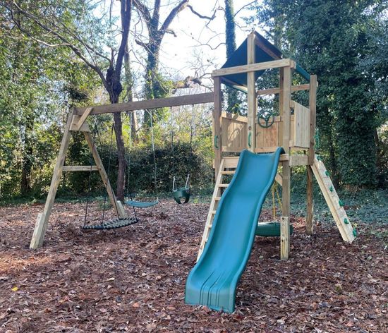 Backyard wooden playset with blue slide, swings, and climbing frame among trees and leaves