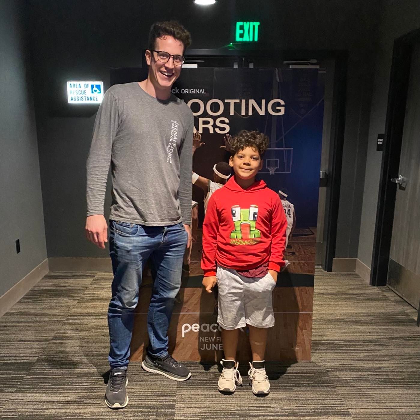 A Big Brother and Little Brother at an event, one in a gray sweater and jeans, the other in a red hoodie and white shorts.