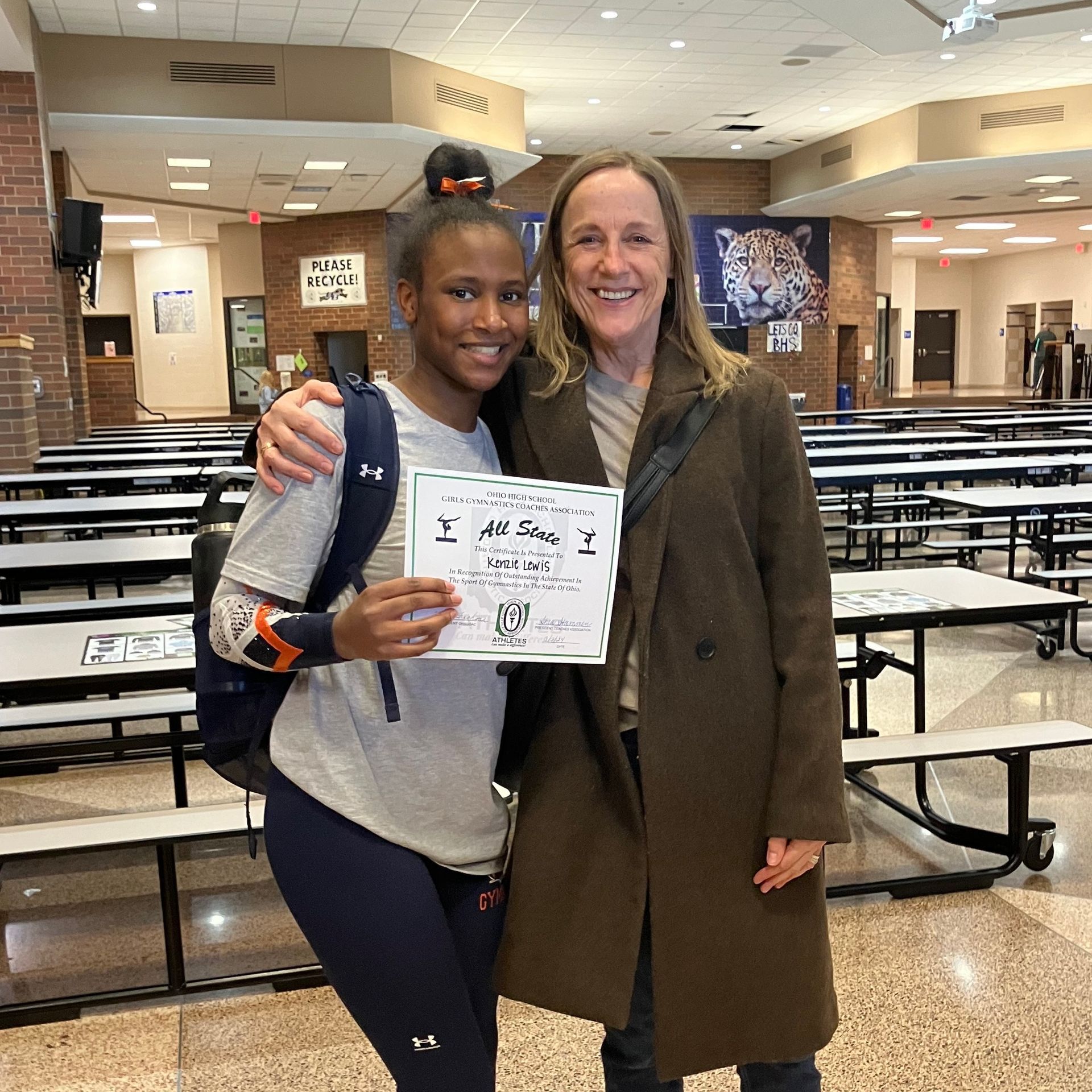 A Big and Little smiling in a cafeteria, holding a certificate and standing arm in arm.
