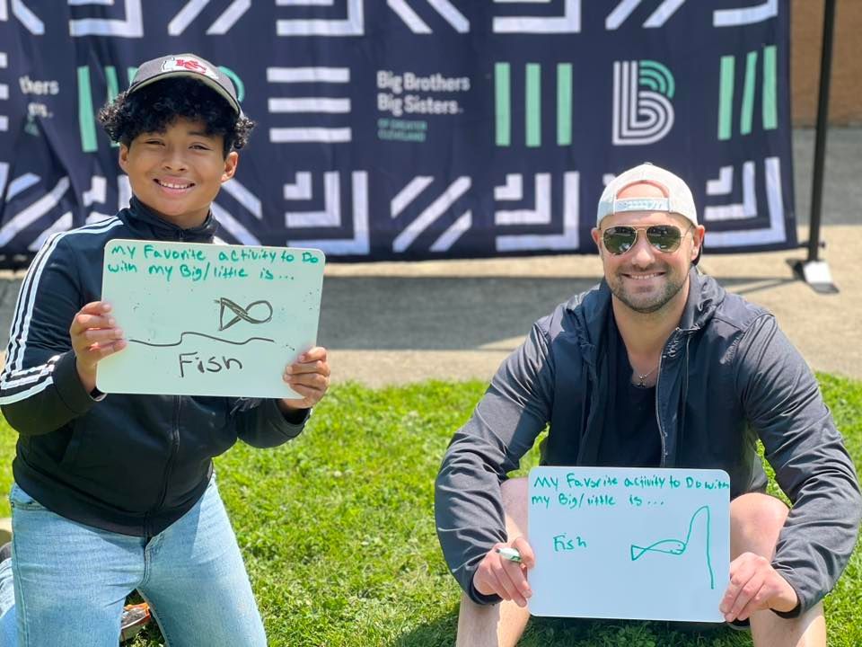 A Big and Little kneel on grass holding handwritten signs in front of a banner.