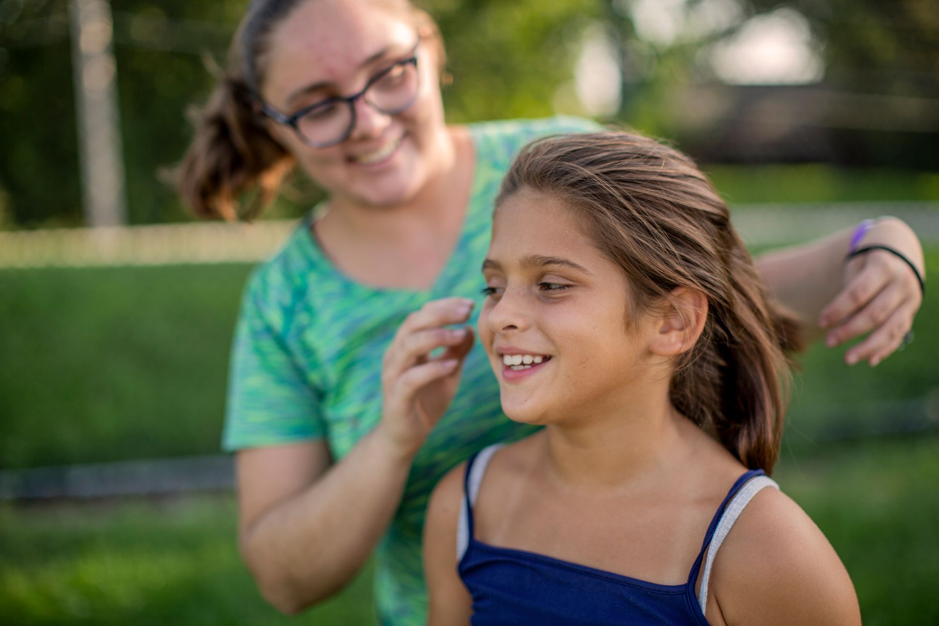 A Big adjusting her smiling Little’s hair outdoors in a green park