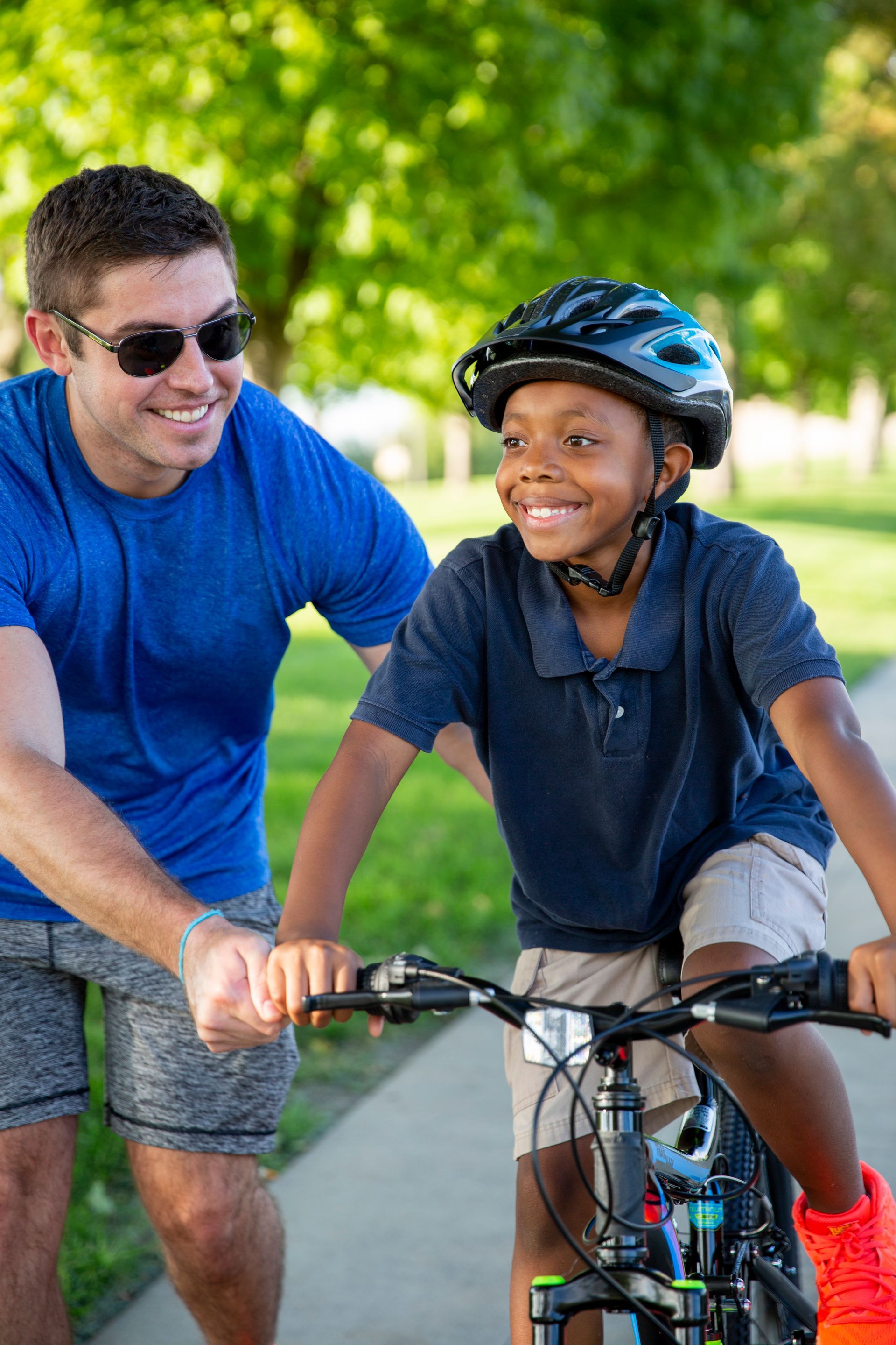 A Big Brother helping his Little Brothers ride a bicycle on a sunny path, both smiling outdoors.