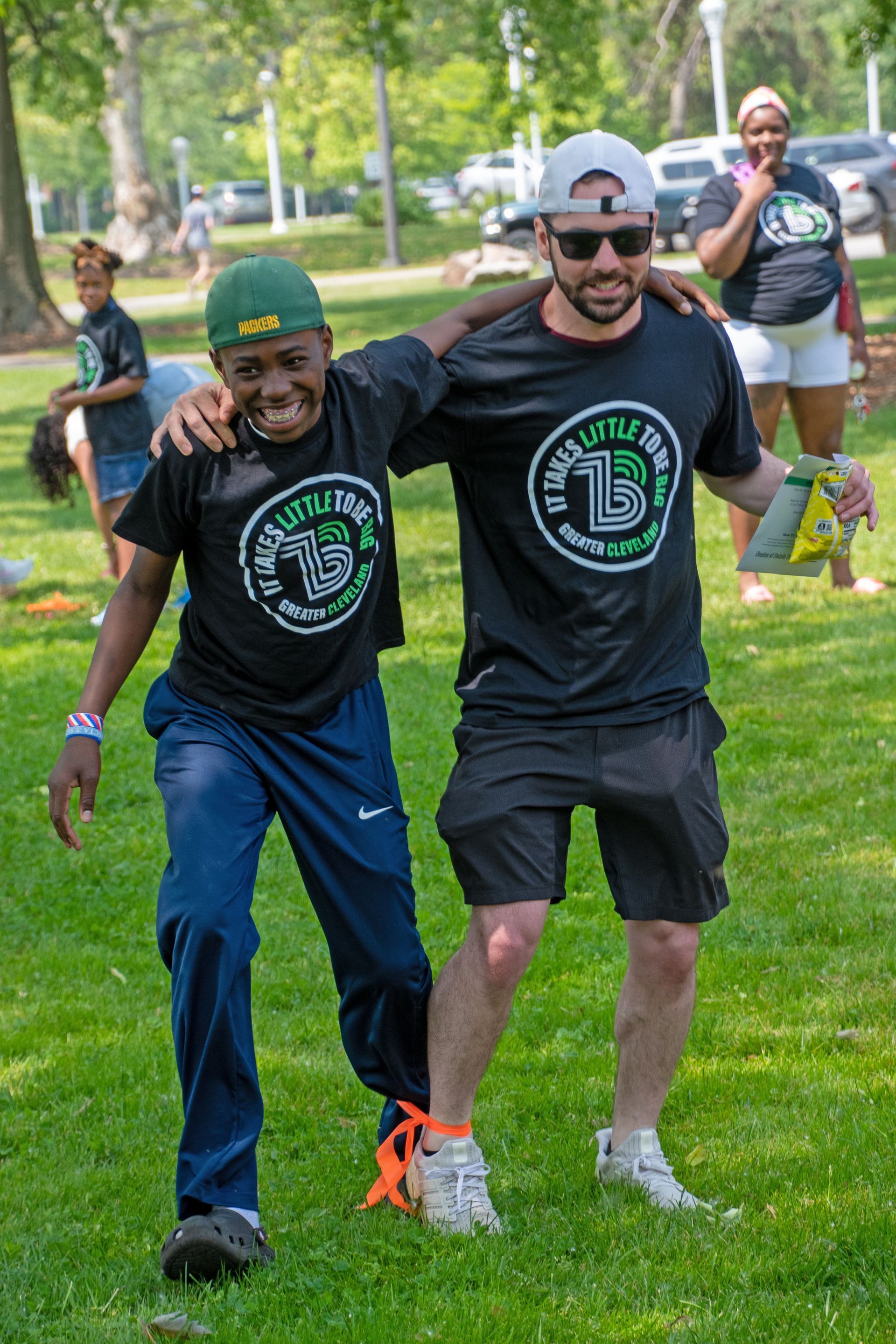 A Big and Little in matching BBBS black shirts are competing in a one-legged race at a BBBBS of Cleveland fundraiser smiling on a sunny day