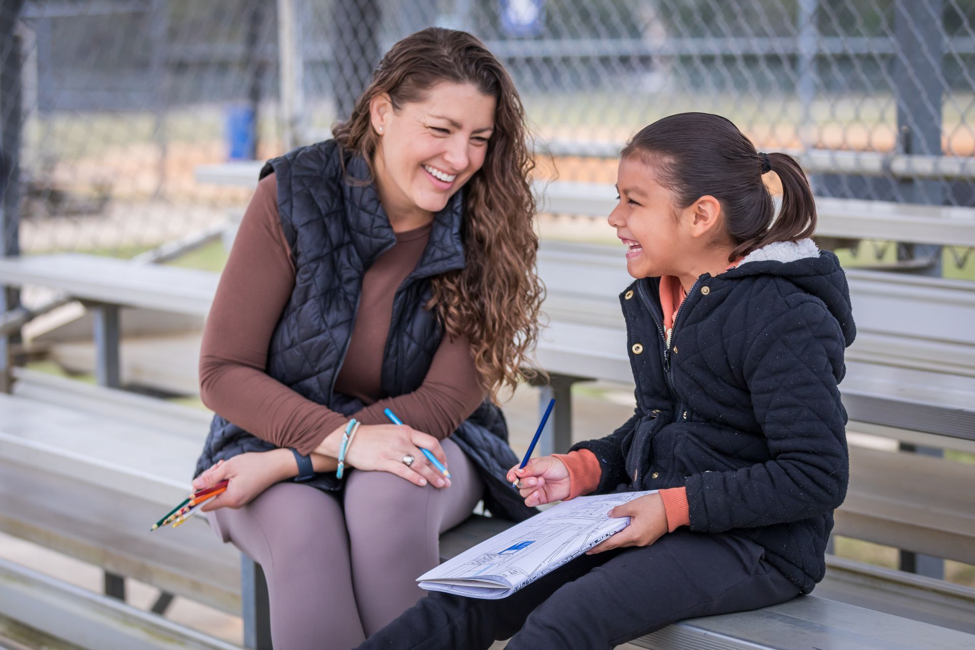 A Big Sister and Little Sister smiling and talking on a bench, holding papers outdoors.