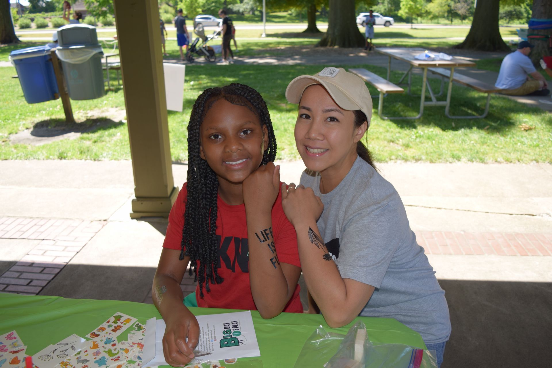 A Big and Little smiling at a picnic table in a park, with a green tablecloth and trees in the background.