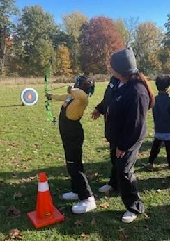 A Little practicing archery outdoors on a grassy field near a target and orange cone.