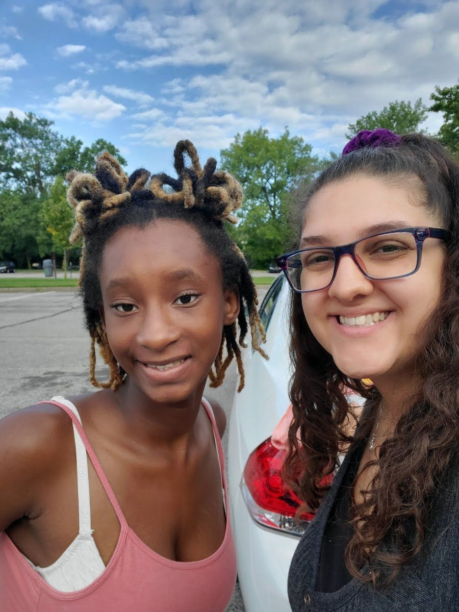 A Big Sister and Little Sister outdoors in a park, with trees and a cloudy sky behind them.