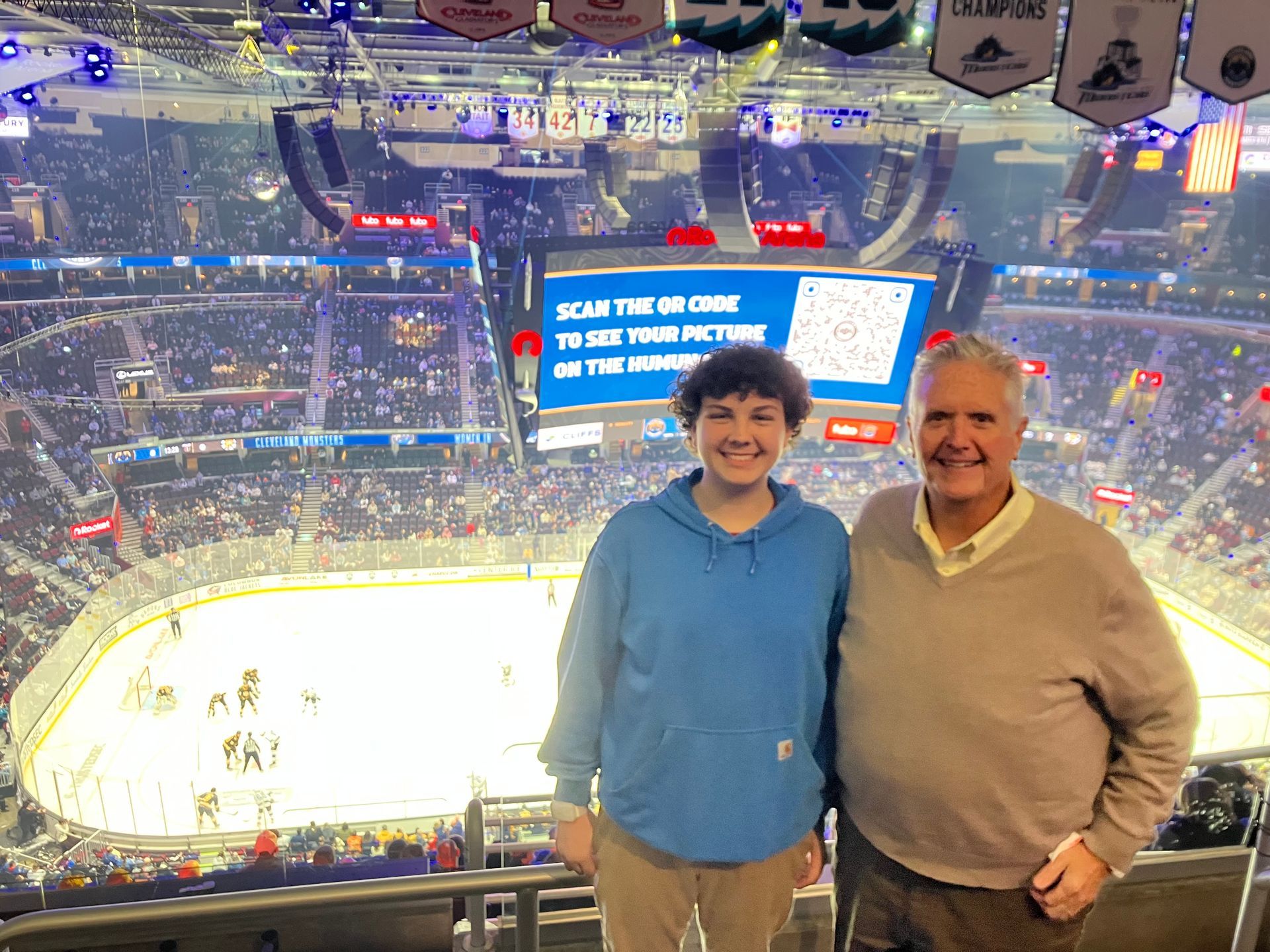 Big Brother with Little Brother smiling in a hockey arena, with the ice rink and a large scoreboard behind them.